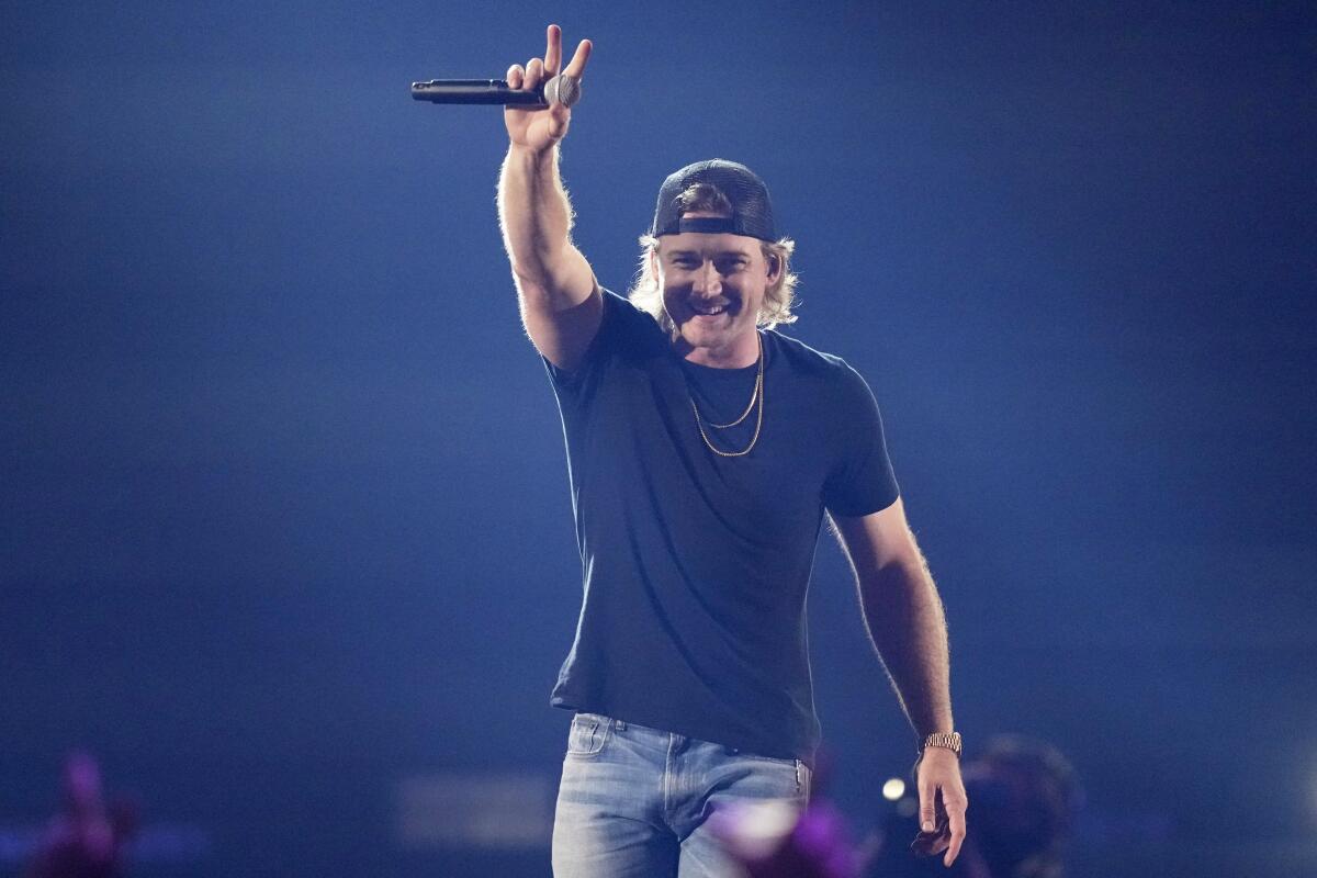 Morgan Wallen holds a microphone and waves to a crowd while wearing a black T-shirt, backward billed hat and jeans.