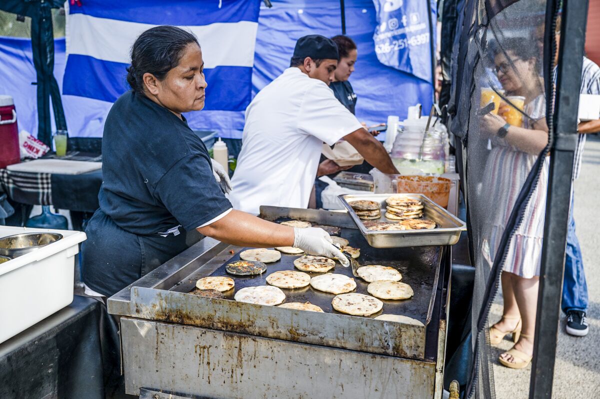 A woman stands at a grill where flat breads are baking; two others prepare dishes at a table beside her.