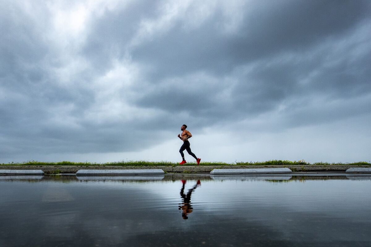 A man jogging is reflected in a pool of calm water. In the background are gray clouds.