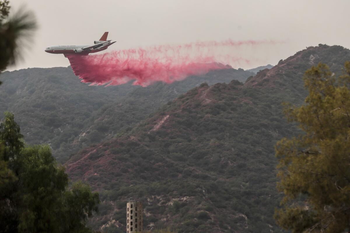 A large jet releases a plume of bright red fire retardant above brush-covered hills