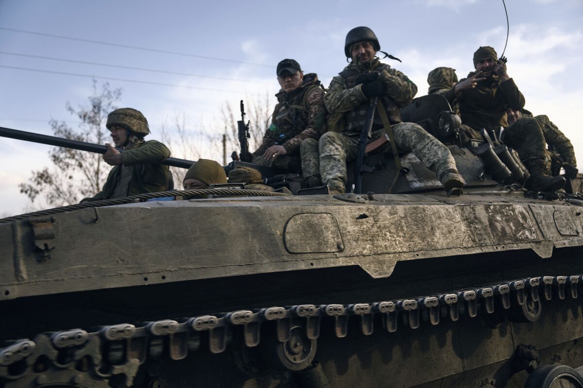 Soldiers in fatigues and helmets ride atop an armored vehicle