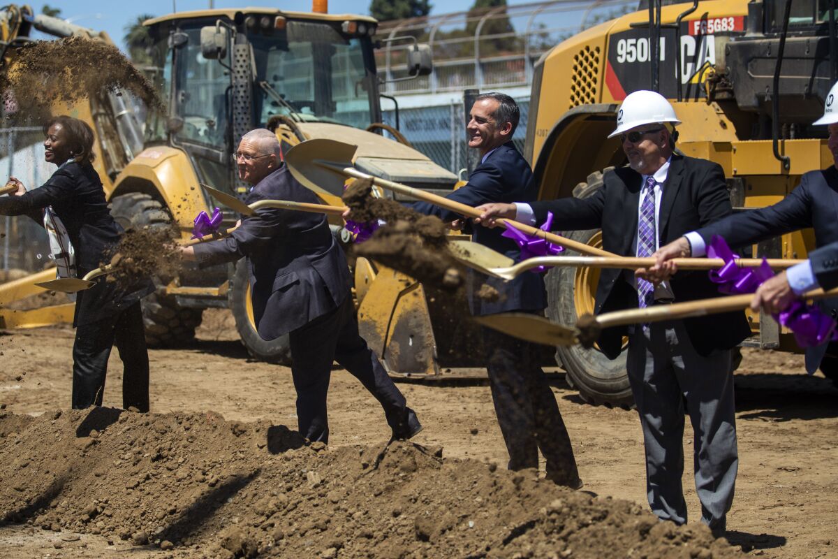 Officials in suits and hardhats wield shovels during a groundbreaking ceremony with heavy machinery in the background