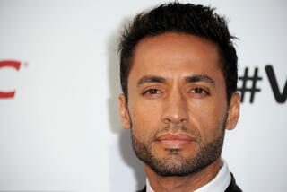 A man with short dark hair and stubble faces the camera in front of a white background