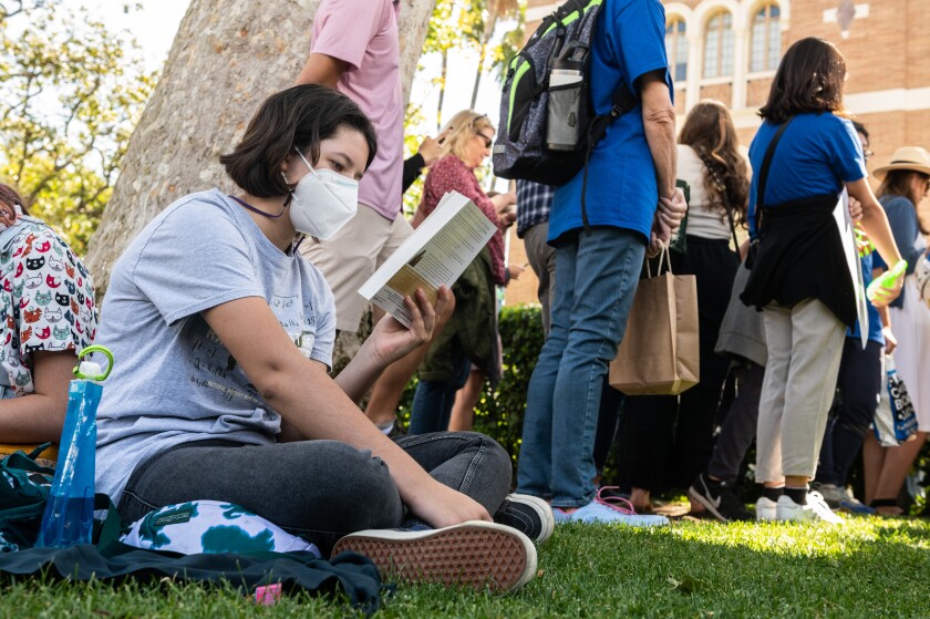 A masked teen reads a book at a book festival.