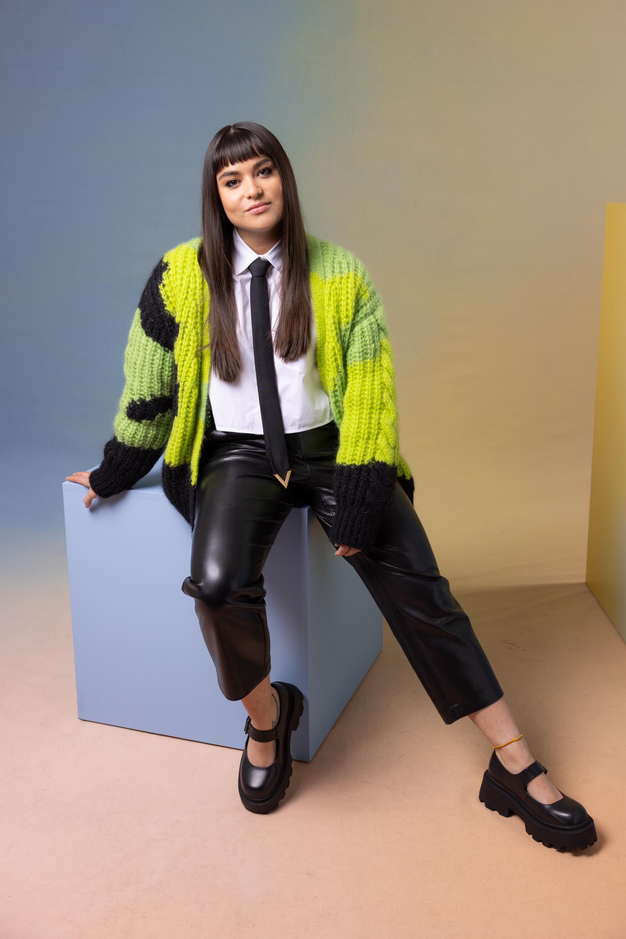 Devery Jacobs wears a black and yellow sweater and black pants while sitting on a cube.