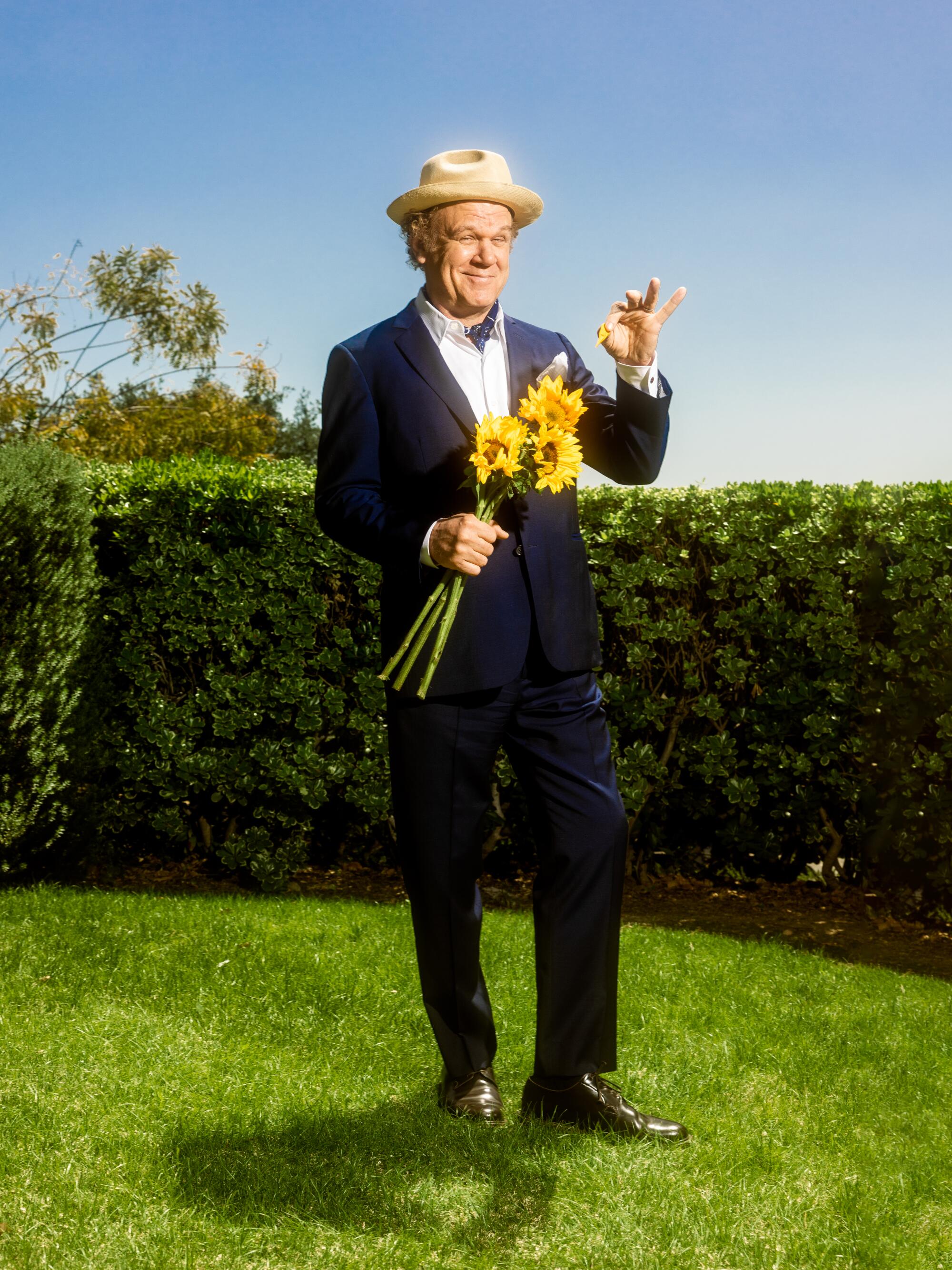 John C. Reilly stands in a grassy patch picking petals off of sunflowers.
