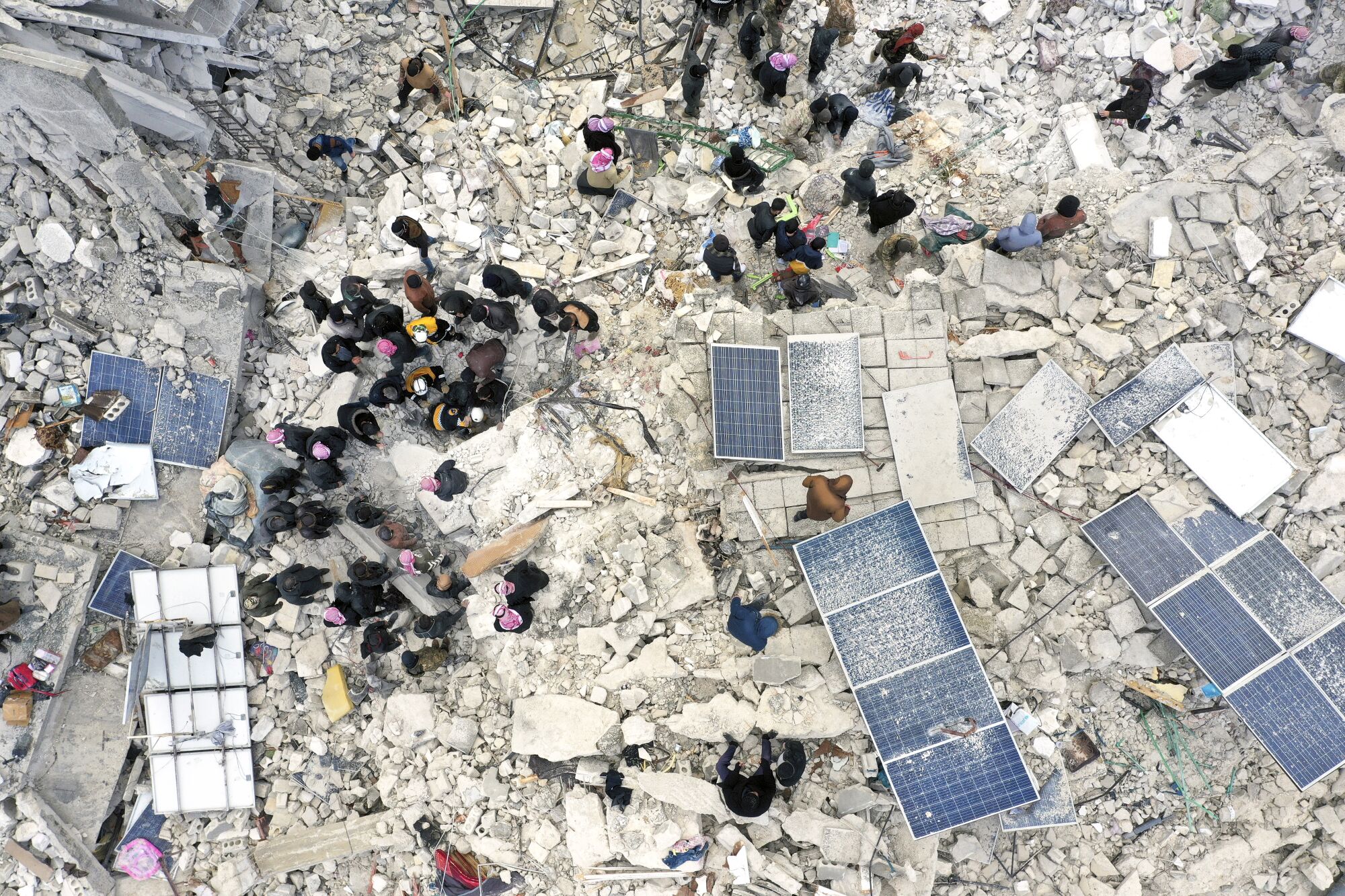 An aerial topdown view of a large area flattened with concrete and debris and mana people walking stop of it.