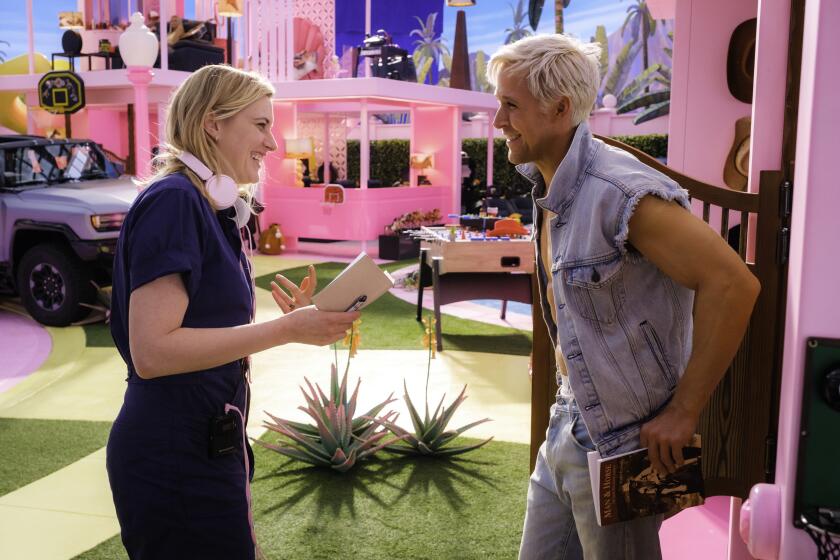 Greta Gerwig, in a dark blue jumpsuit, smiles and chats with Ryan Gosling, in a jean jacket, while on the set of Barbie