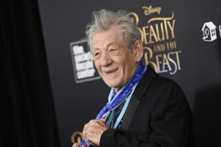Ian McKellen in a black blazer, holding a blue neck scarf around his neck and posing against a black backdrop