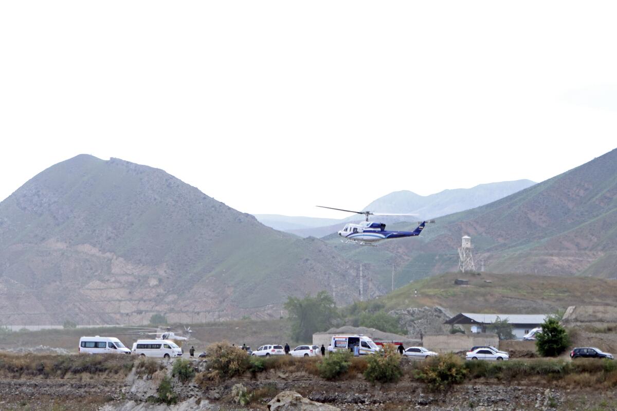 A helicopter hovers over several vehicles with mountains in the background.