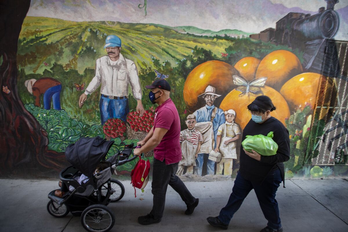 Pedestrians, one pushing a stroller, wear masks as they pass a mural of farmworkers