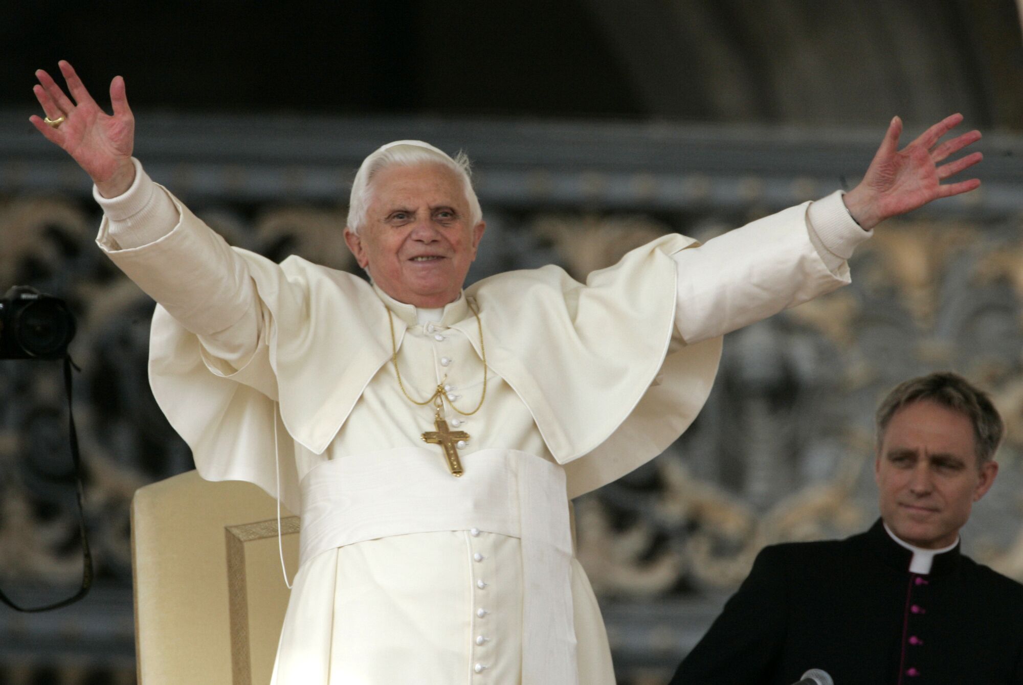 پاپ بندیکت شانزدهم در حالی که مرد دیگری به او نگاه می کند، دستانش را بالا می برد.