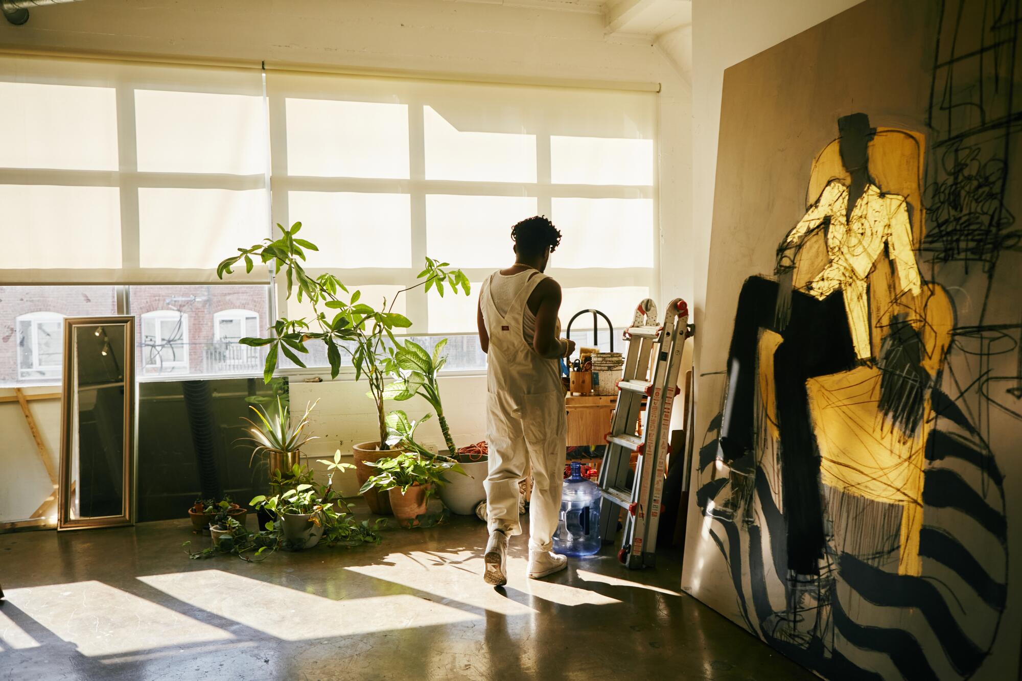 An artist in his studio, with lots of windows, stands before a painting of a seated woman in a black jacket 