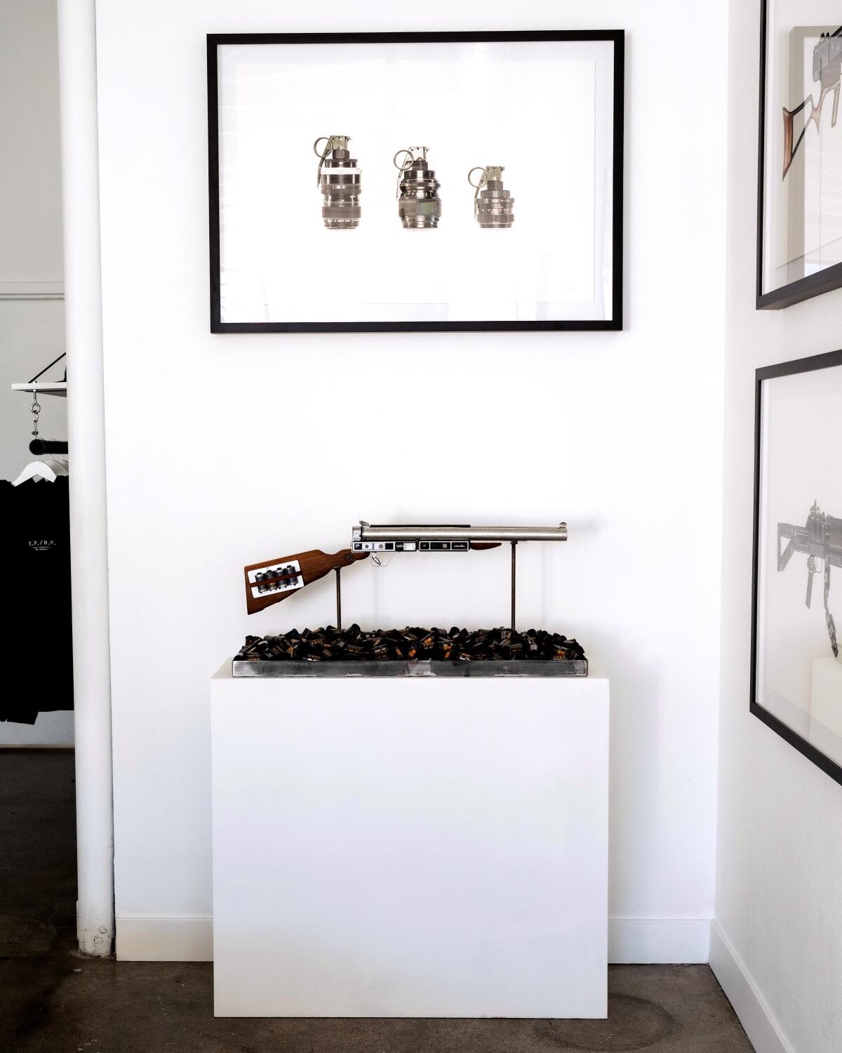 A gallery space with an art sculpture of a gun on a pedestal with a picture of grenades on the wall.