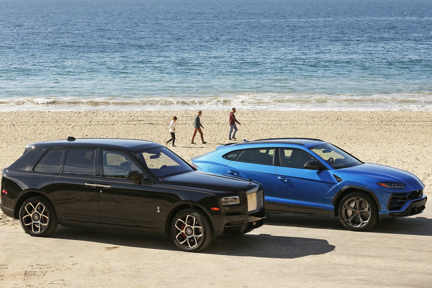 The Rolls-Royce Cullinan Black Badge, in black, and the Lamborghini Urus, in blue, on a beach.