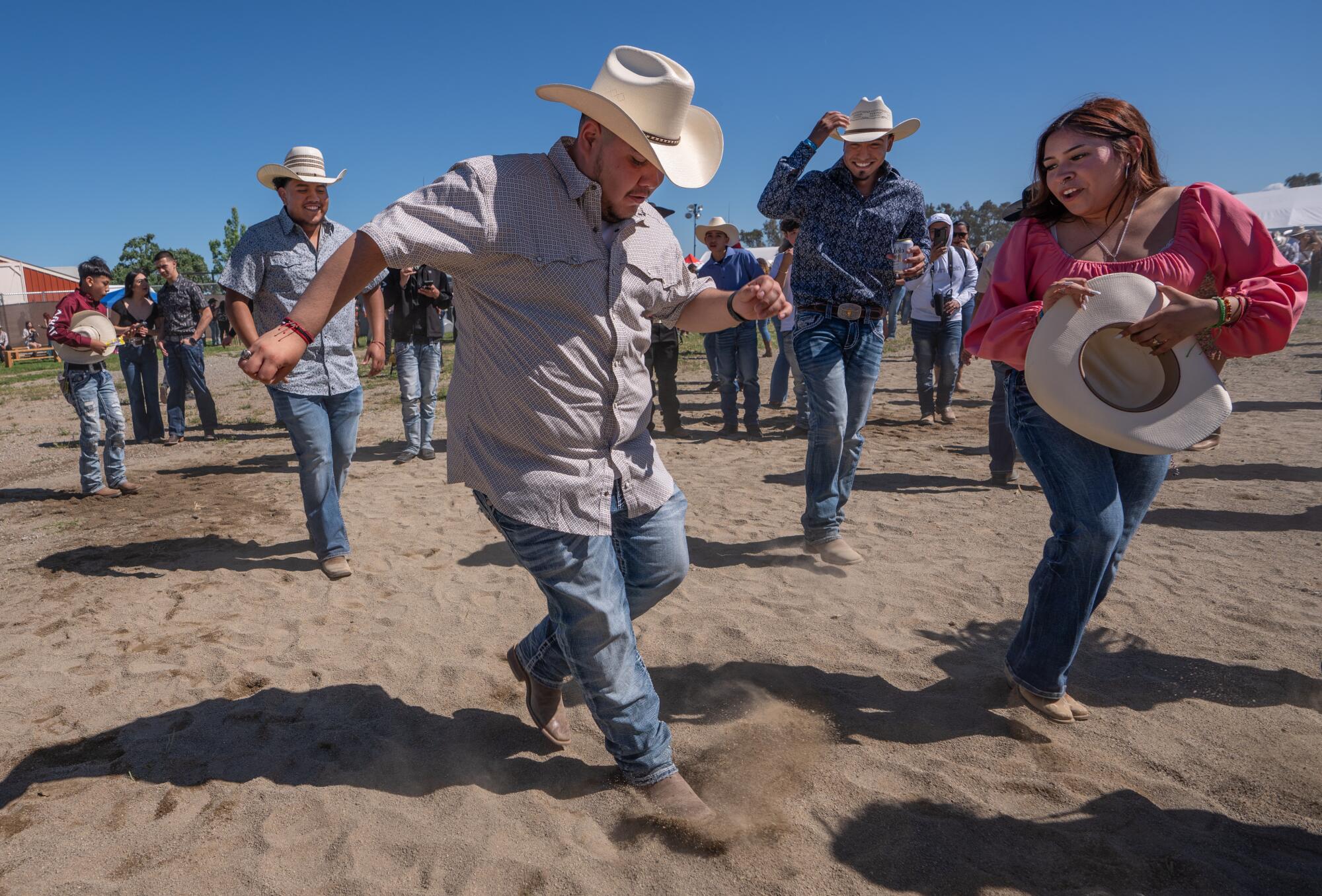 Attendees dance in cowboy hats and jeans dance in the dirt at a music festival.
