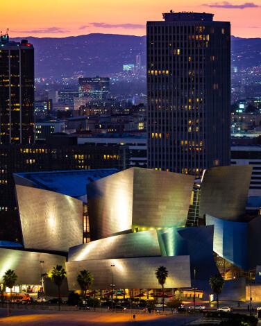 Walt Disney Concert Hall's silvery structures illuminated at twilight, with skyscrapers and hills in the distance