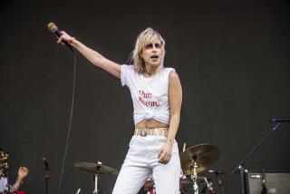 A blond woman wearing rainbow face paint and a white outfit holding a microphone in the air on a stage