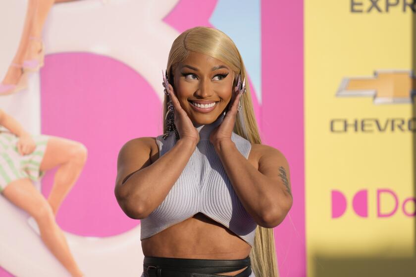 Nicki Minaj in a matching blue-gray top and skirt holding her hands to her face and smiling in front of a Barbie poster