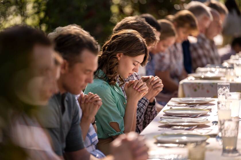 A woman at a long dining table bows her head in prayer 