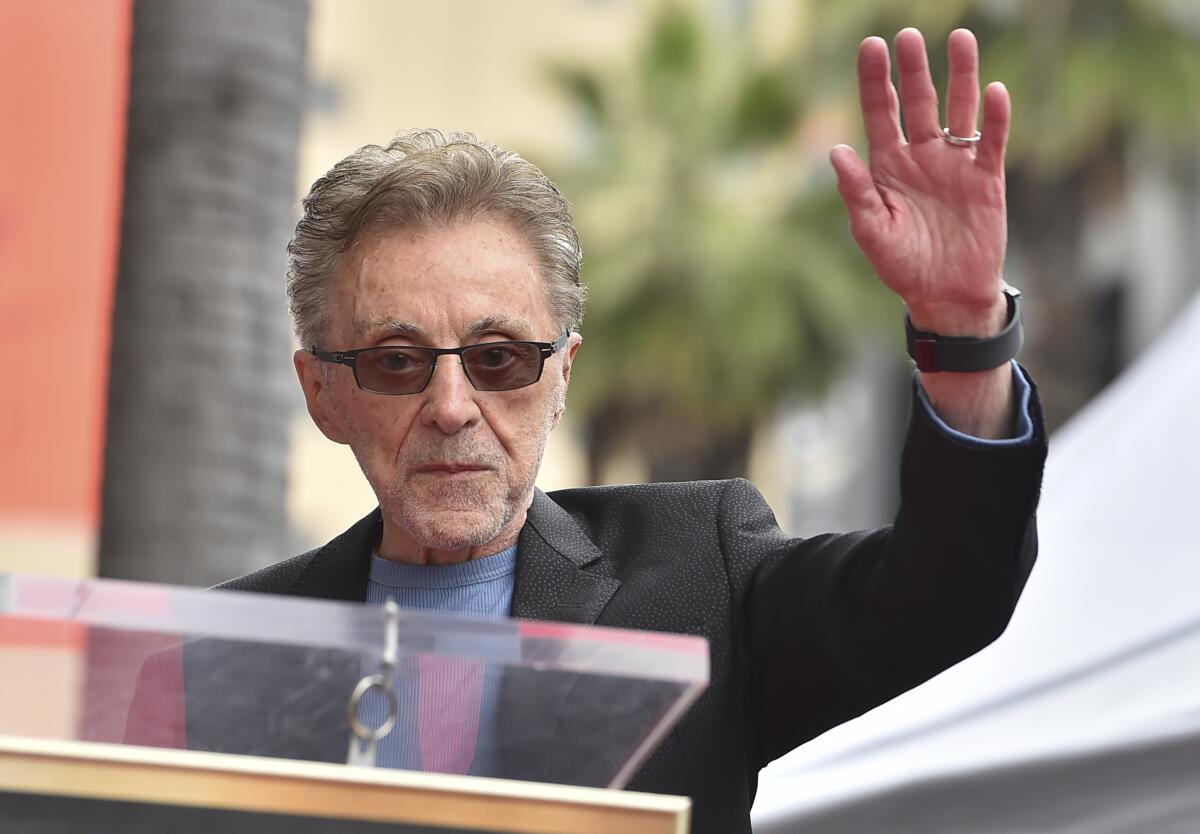 Frankie Valli in sunglasses, a blue shirt and a dark suit jacket holding up his left hand as he approaches a lectern