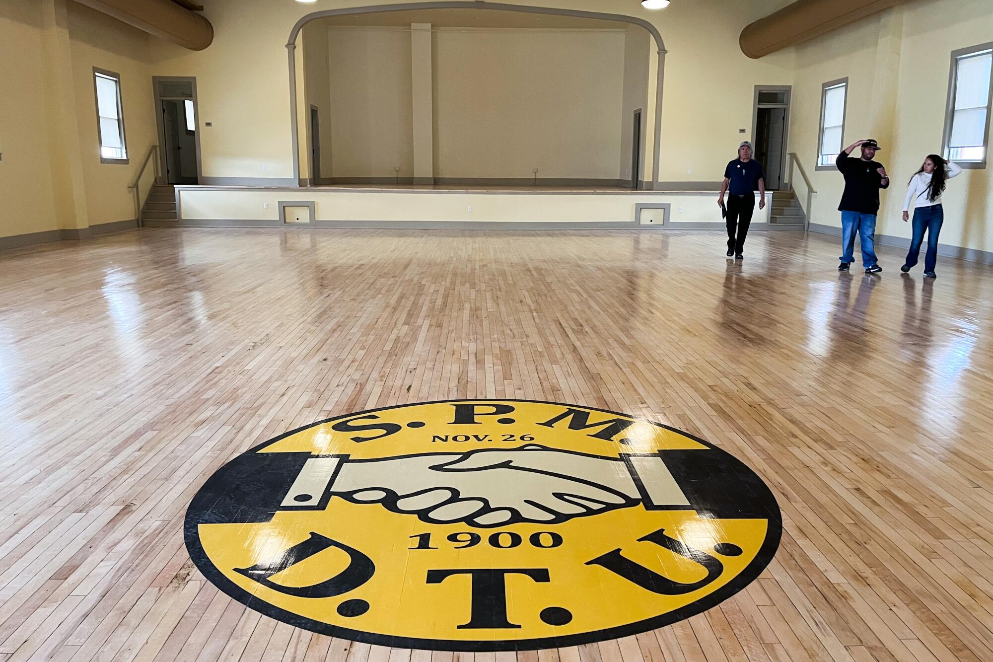 A round yellow logo on a hardwood floor reads "S.P.M.D.T.U." in black letters, bracketing two clasped hands.