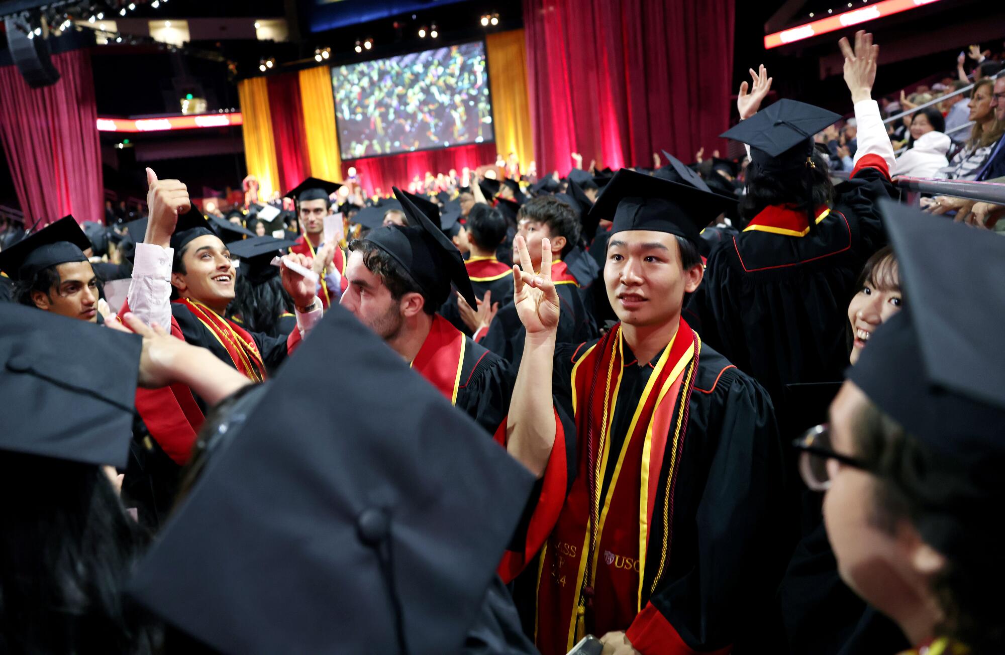 A crowd of USC graduates, one of them giving the "Fight" hand gesture, in black robes and mortarboard hats