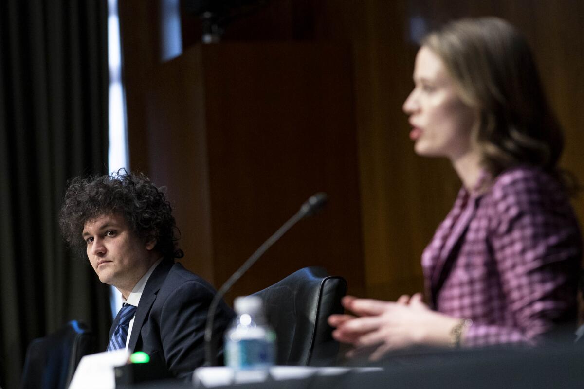 Sam Bankman-Fried sits and listens while a woman speaks at a microphone