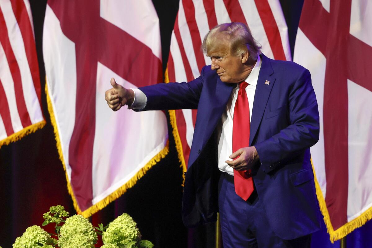 Donald Trump gives a thumbs-up from a stage with flags in the background