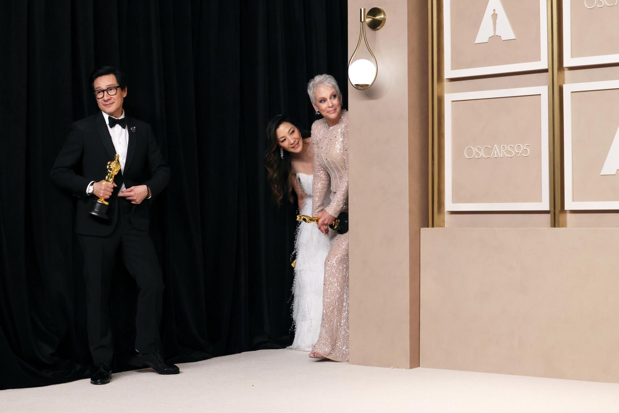 Three people in evening wear - a man stands at left while two women peek in from behind a wall with the writing "Oscars." 