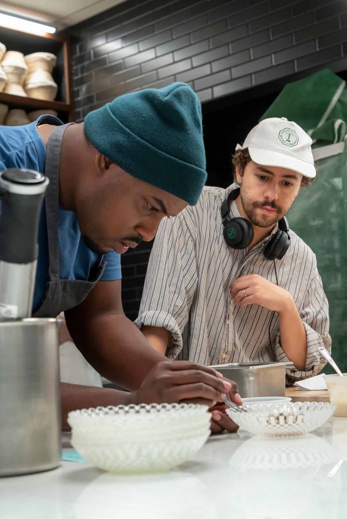 Lionel Boyce and Ramy Youssef, who has headphones around his neck, standing in a kitchen