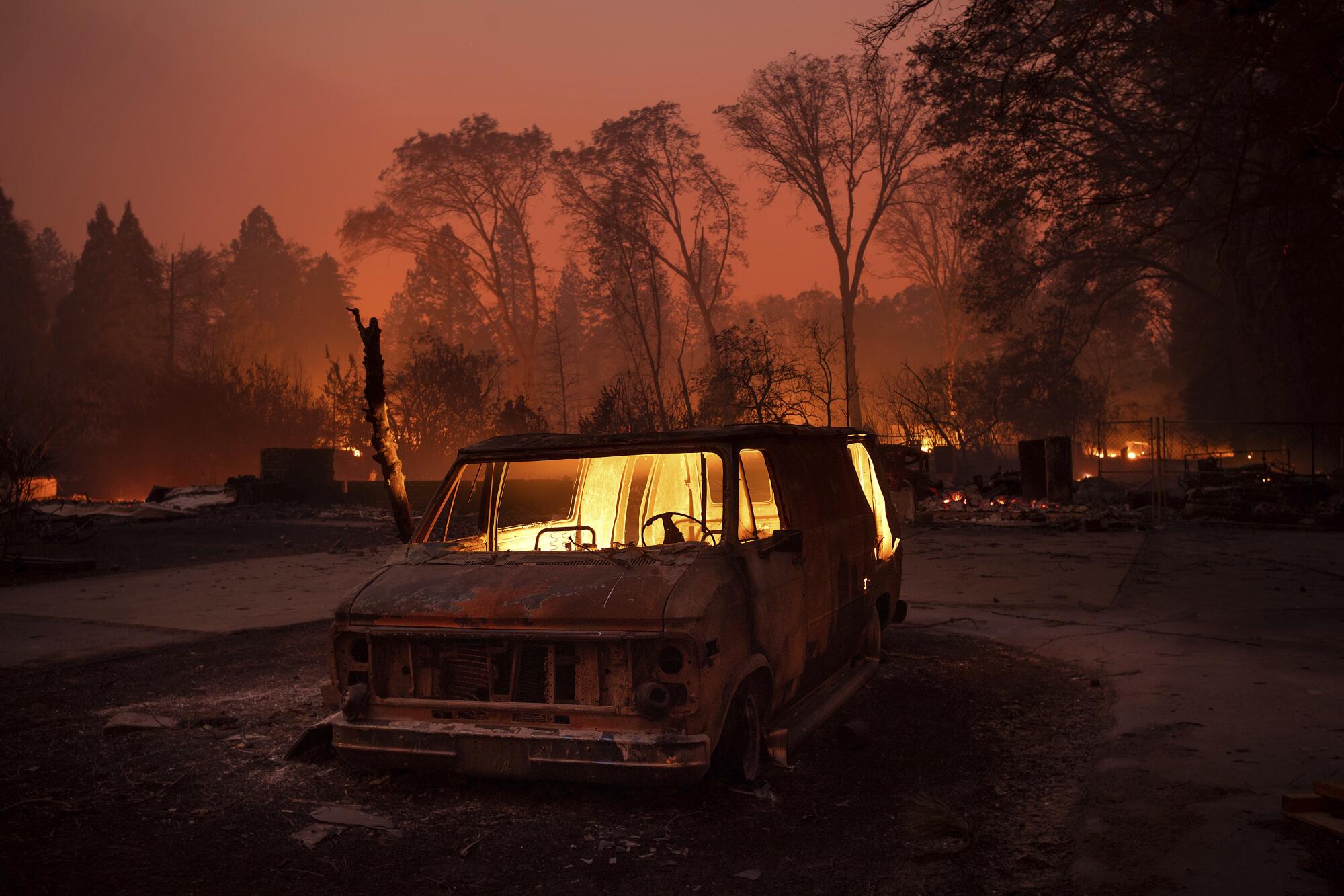 Flames burn inside a van during the Camp fire as trees are in the background and the sky is orange. 