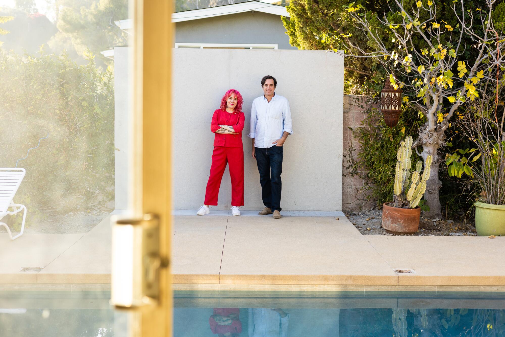 A woman in red and a man in a white shirt and black pants stand in front of a pool in his back yard.