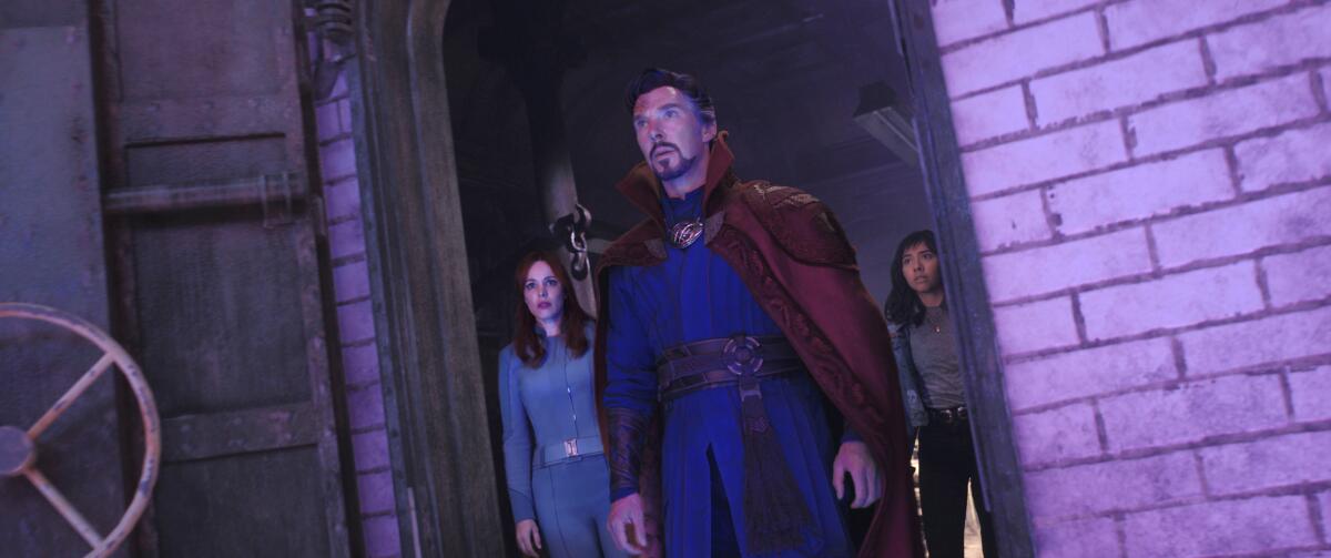 Two women and a man in a superhero costume stand in an open doorway looking up.