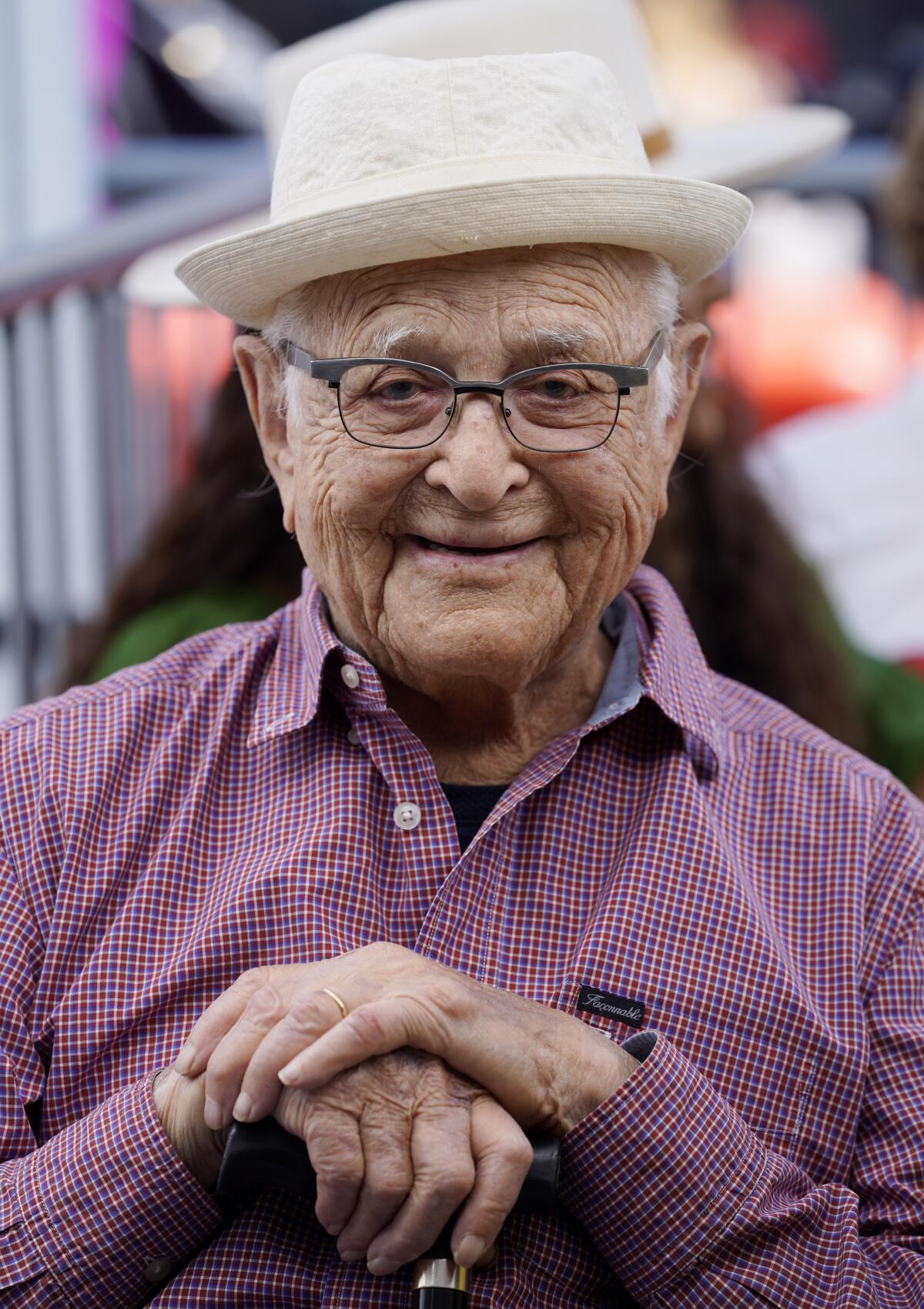 A smiling, older man with glasses, in white hat and red checkered shirt, with hands together 