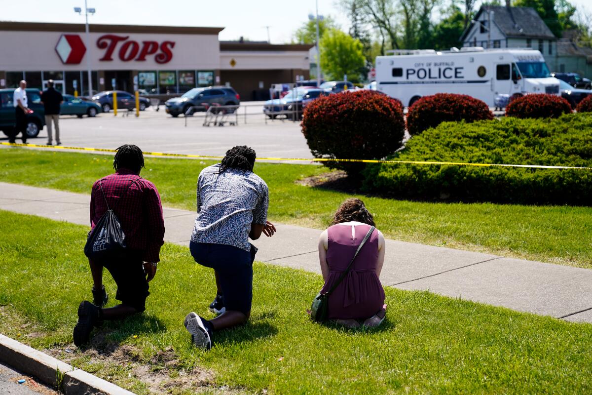 Three people kneeling in grass along a sidewalk outside a Tops supermarket.