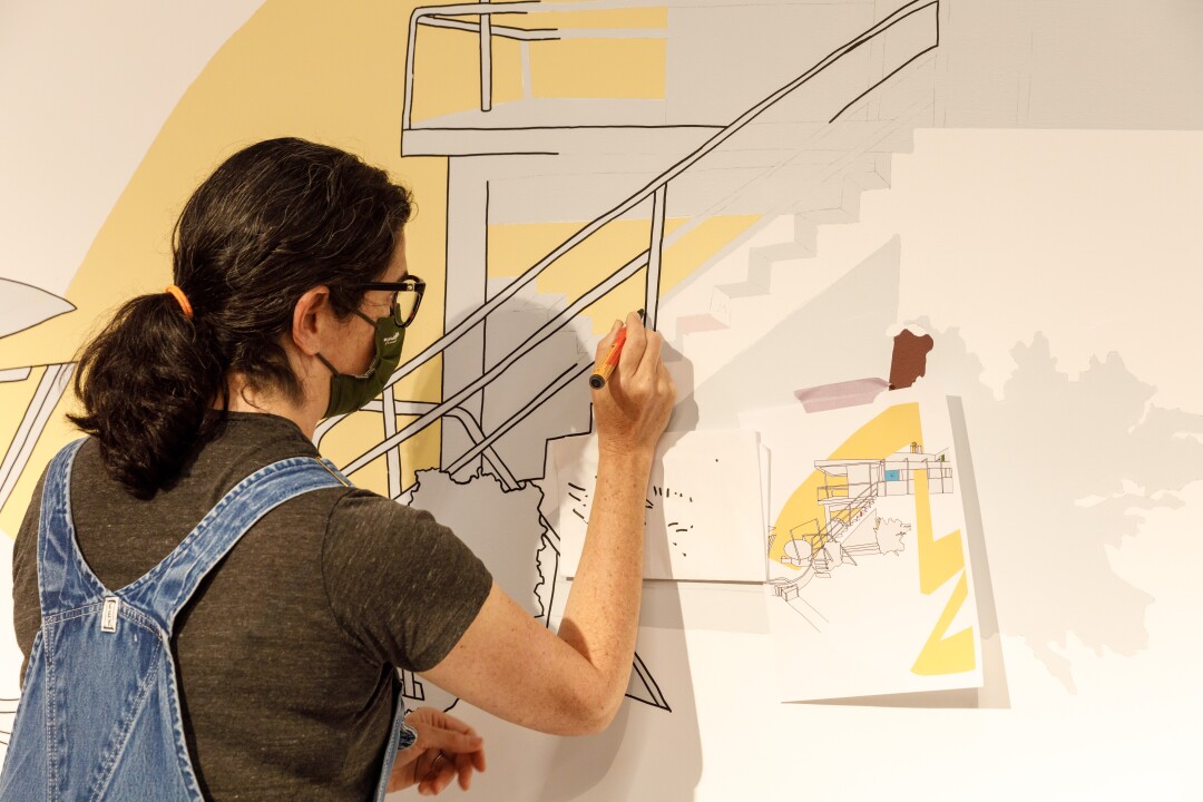 Kim Schoenstadt, in overalls, is seen drawing a staircase into an architectural drawing on a wall