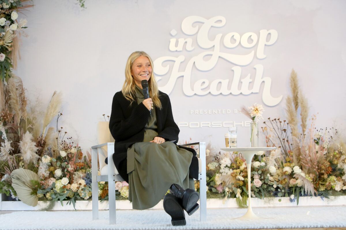 Gwyneth Paltrow, in a loose olive dress, sits in a chair on a dais before a large sign that reads "In Goop Health."