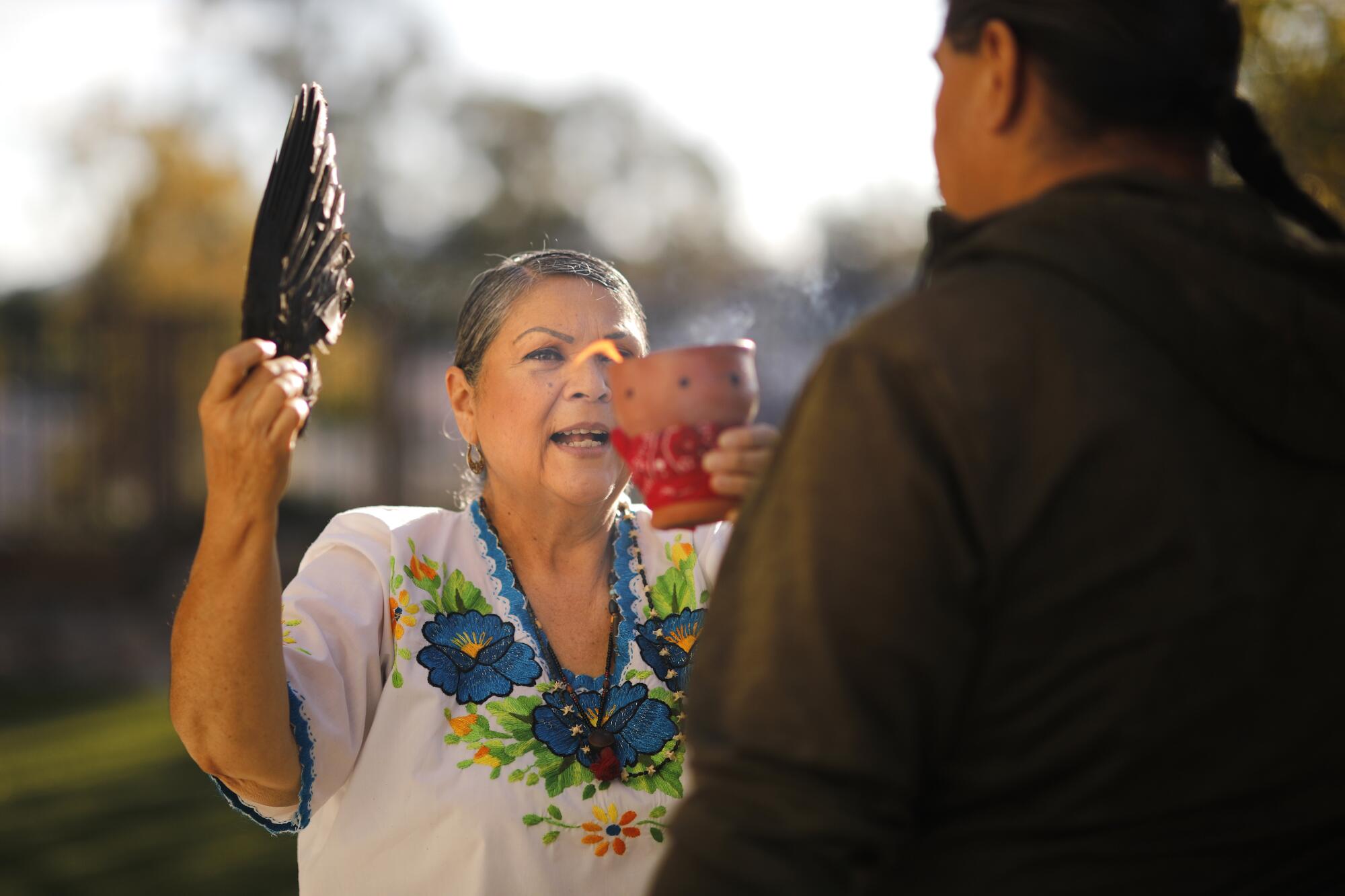 A curandera wearing a floral shirt wafts smoke toward a client.