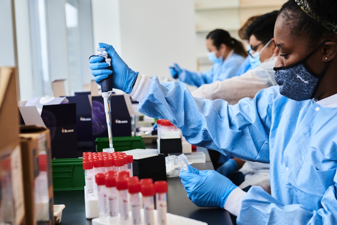 Workers in blue gloves and lab suits hold hand pipettes over tubes with red caps 