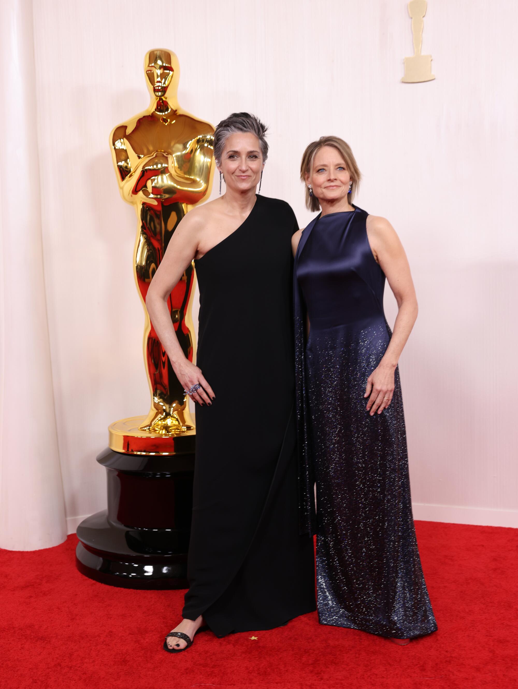 Alexandra Hedison, in a black one-shoulder gown, and Jodie Foster, in a navy blue gown, pose for cameras. 