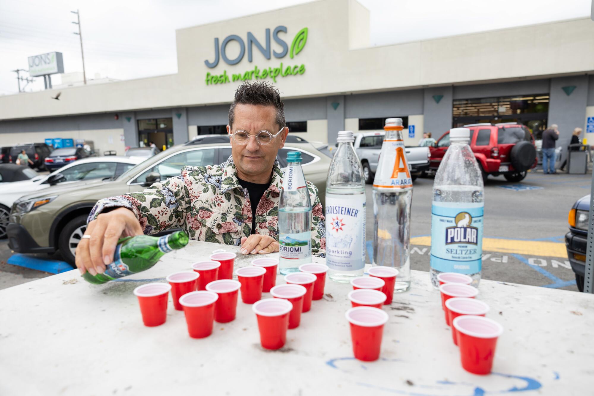A man pouring water into tiny red Solo cups in the parking lot of a grocery store.