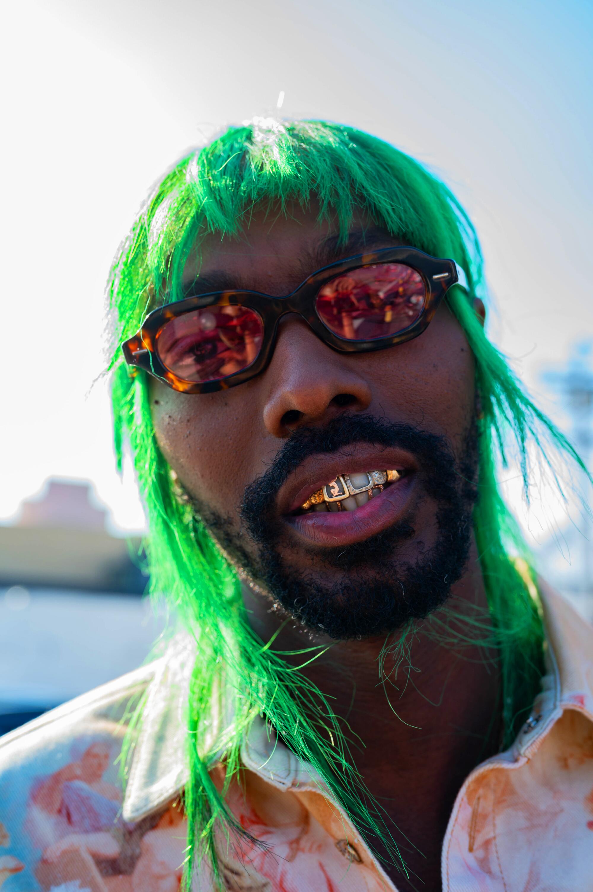 A person, wearing green hair and sunglasses, smiles.
