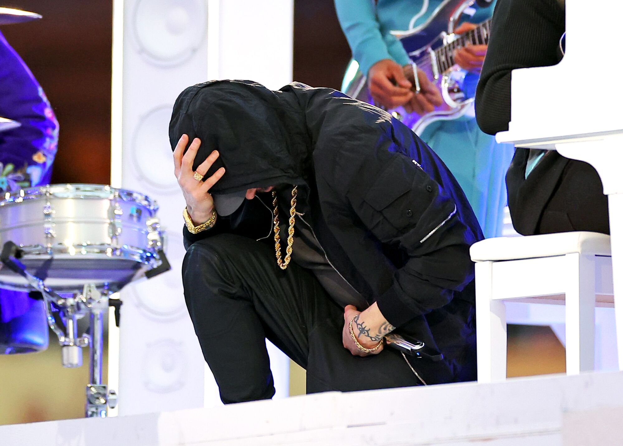 A man in a black hoodie and black pants kneels onstage.