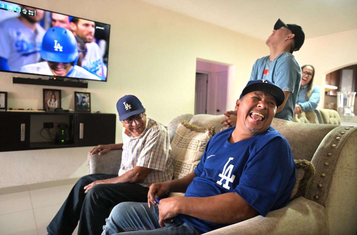 People laugh inside a living room as they watch a baseball game on TV.
