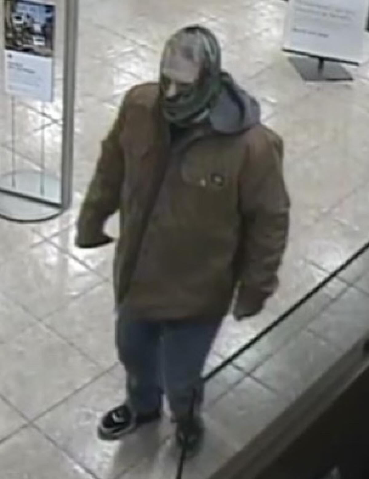 A person is shown wearing a green neck gaiter, a tan jacket, blue jeans and black sneakers.