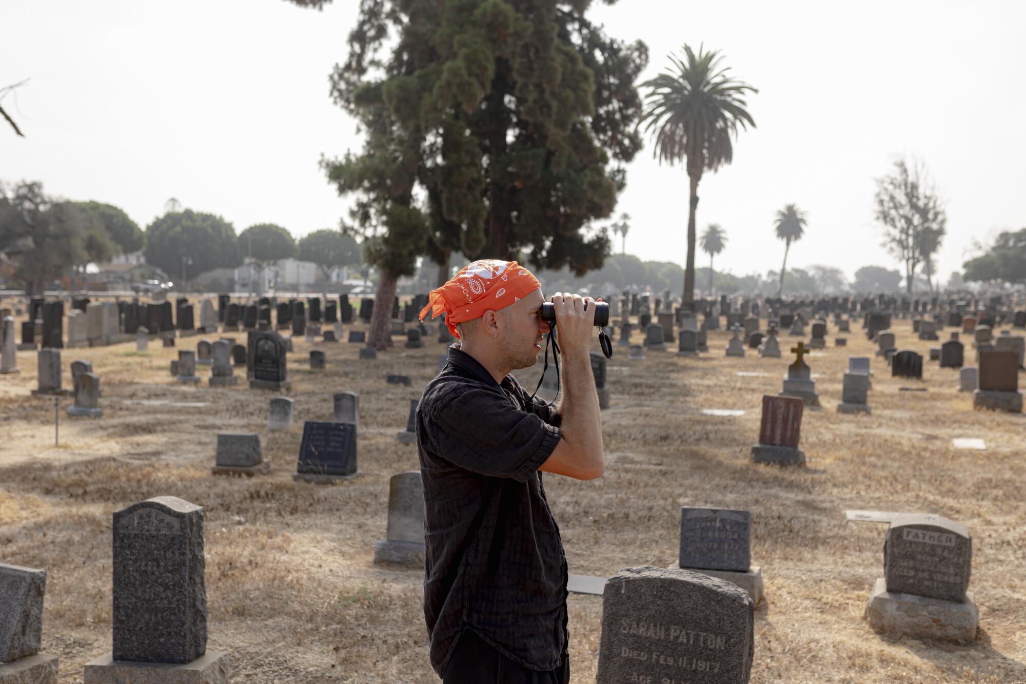 A man wearing a bandana scans a cemetery through binoculars