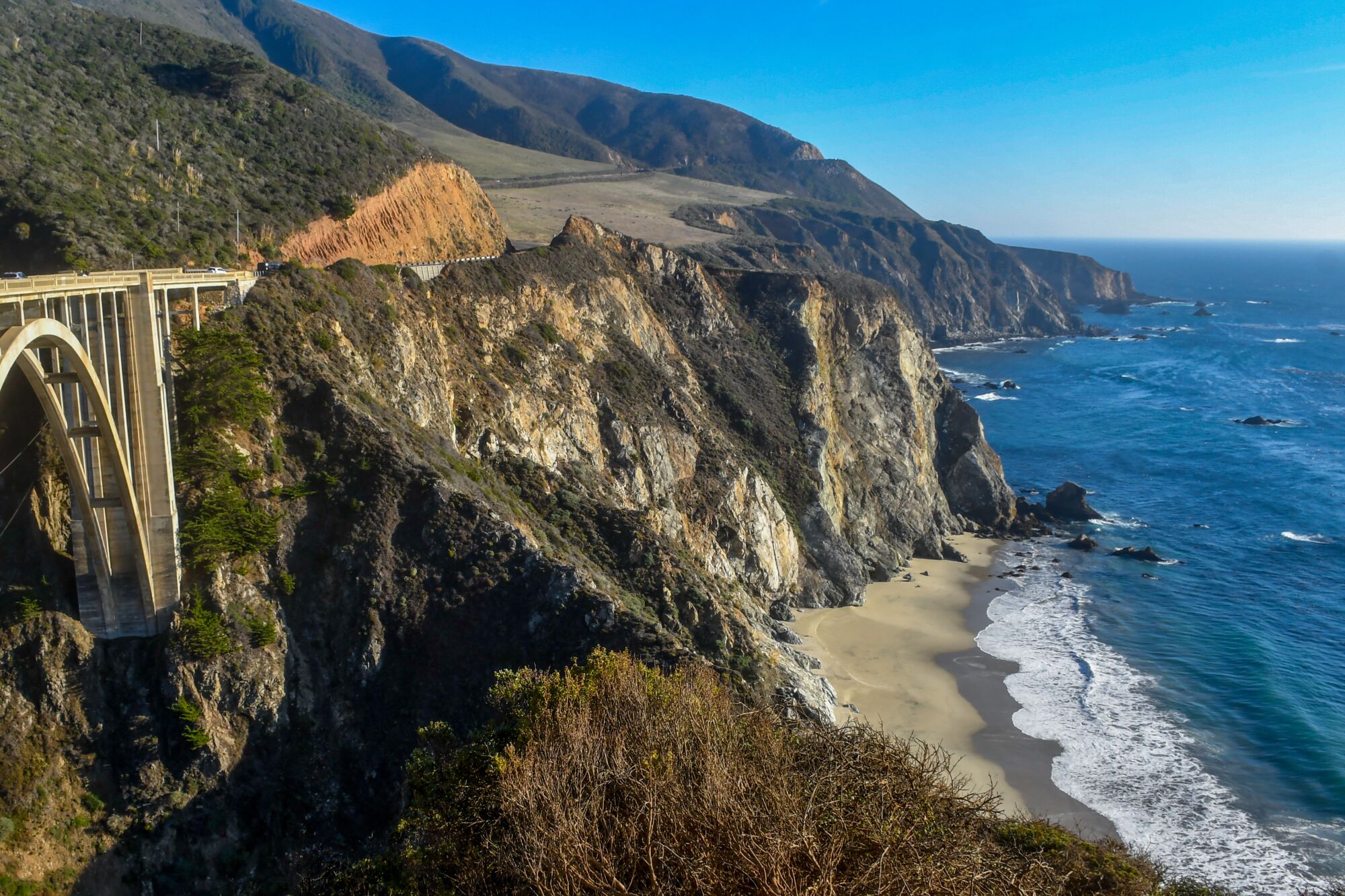 A bridge to the left of a cliffside beach scene.