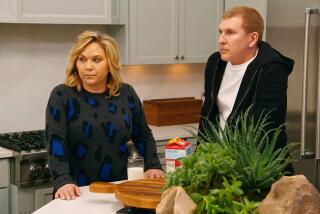 A husband and wife on a reality TV show stand in a kitchen looking concerned