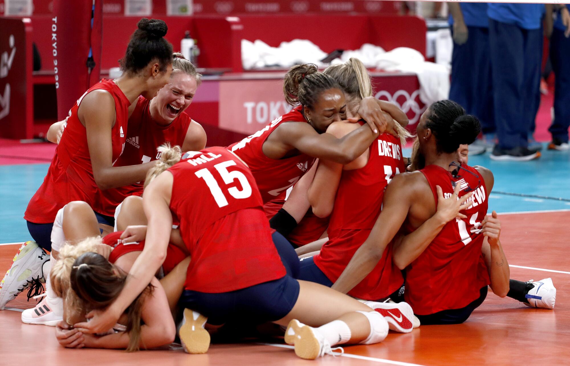 Smiling women form a heap on the floor of an indoor volleyball court.